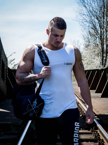 Fitnessmodel trägt das Gym Generation Fitness Tank Top 'Warrior' in Weiß, kombiniert mit Warrior-Jogginghose und Gym Generation Sporttasche, auf einer Brücke.