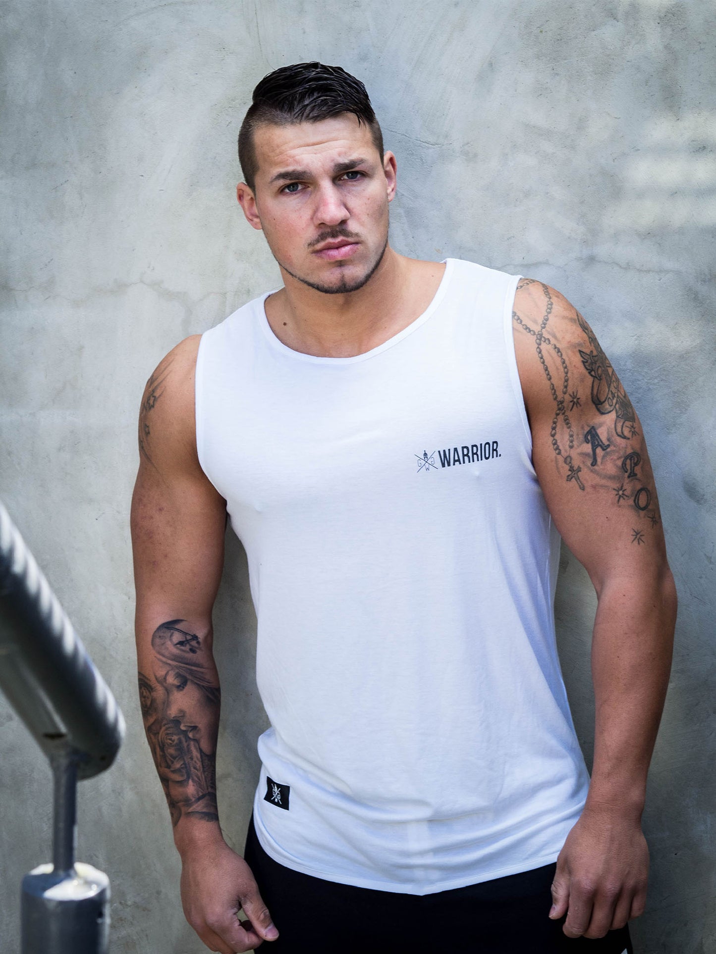 Fitnessmodel trägt das Gym Generation Fitness Tank Top 'Warrior' in Weiß, posiert vor einer Betonwand im Outdoor-Bereich.