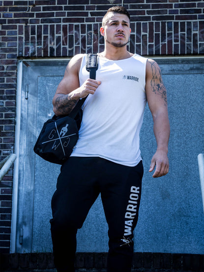 Fitnessmodel trägt das Gym Generation Fitness Tank Top 'Warrior' in Weiß, kombiniert mit Warrior-Jogginghose und Gym Generation Sporttasche, posiert vor einer Backsteinwand.