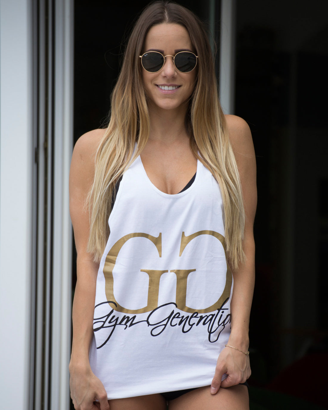 Weisses Gym Generation Stringer Tank Top mit goldenem Logo, getragen von Model Vanessa Mariposa mit langen blonden Haaren und Sonnenbrille.