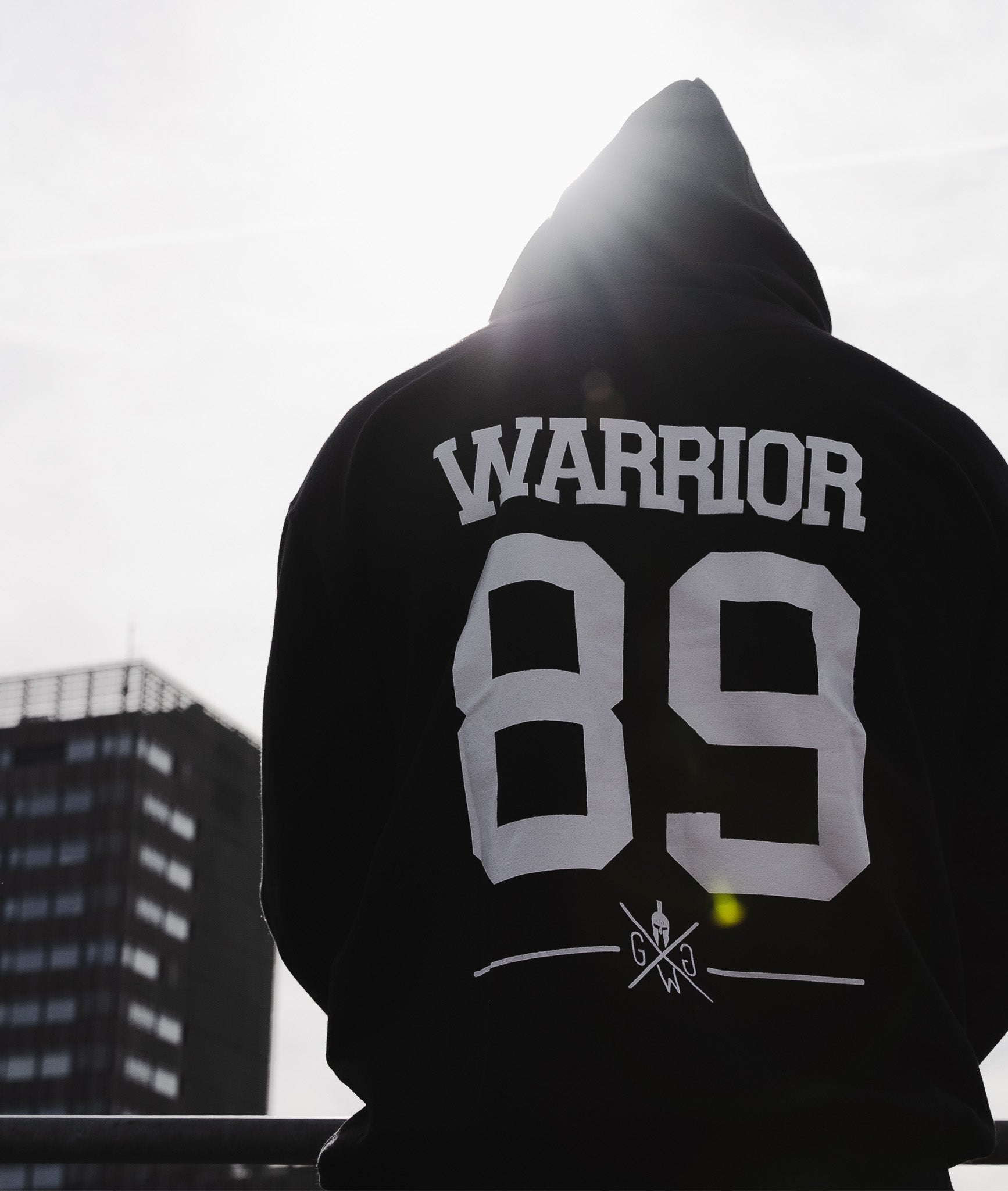 Rückansicht eines Gym Generation Hoodies in Schwarz mit 'Warrior 89'-Aufdruck, fotografiert vor urbaner Skyline im Gegenlicht.