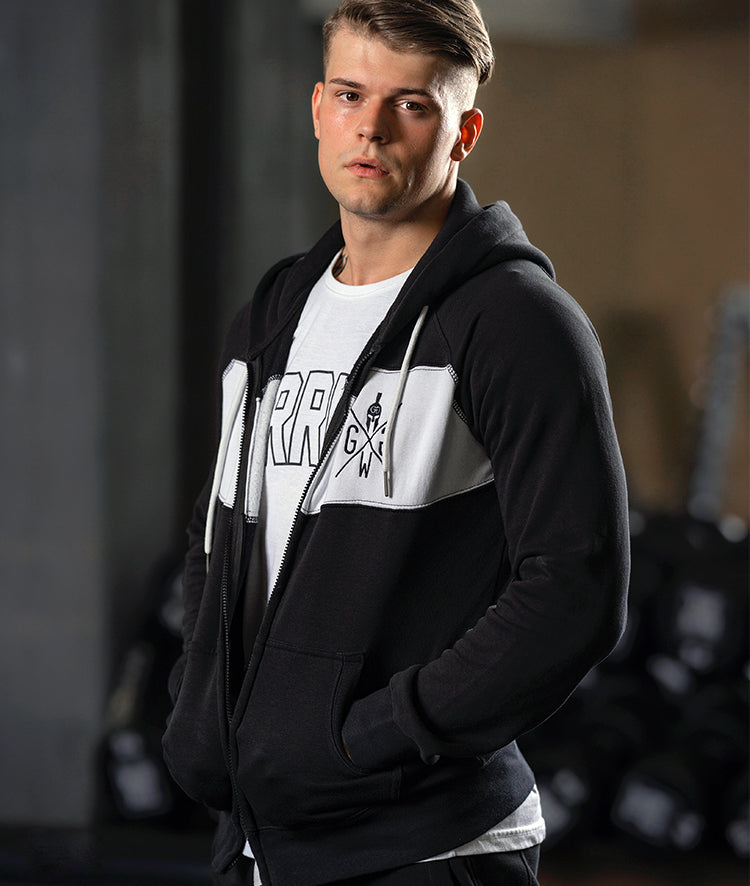 Muskulöser Mann in einer schwarzen Gym Generation Hoodie-Jacke und T-Shirt vor einer Fitnessstudio-Kulisse mit Gewichten im Hintergrund.