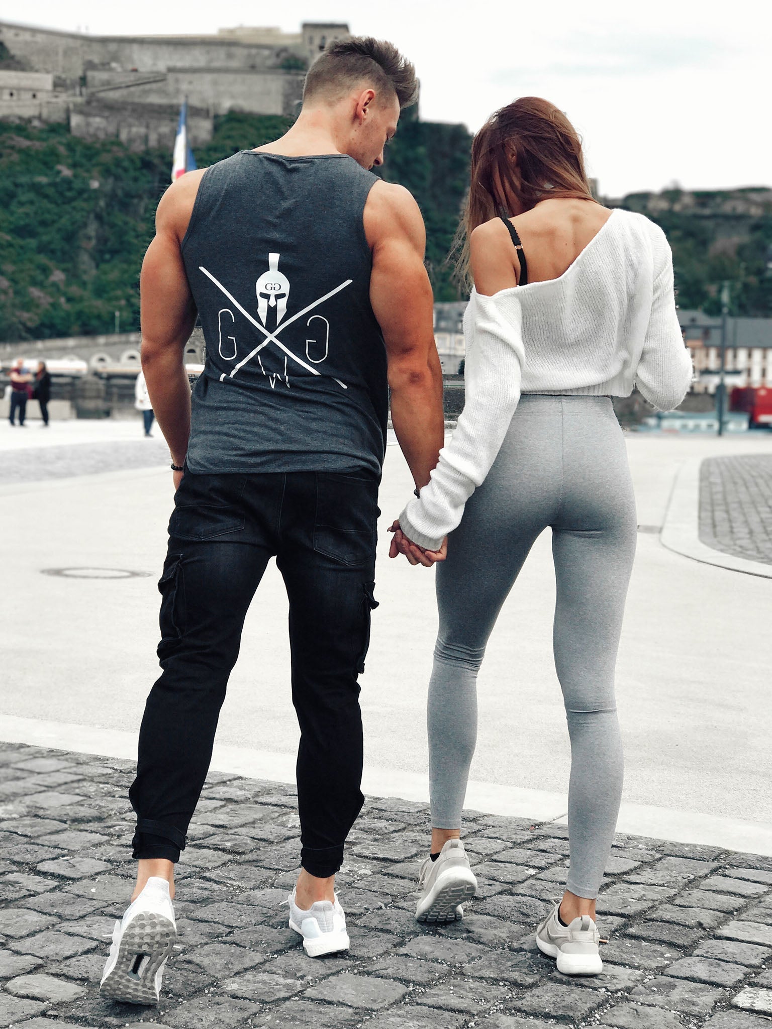 Paar in Gym Generation Kleidung: Eine Person trägt das Gym Warriors Tank Top, während die andere Fitness-Leggings trägt, in einer urbanen Outdoor-Umgebung.