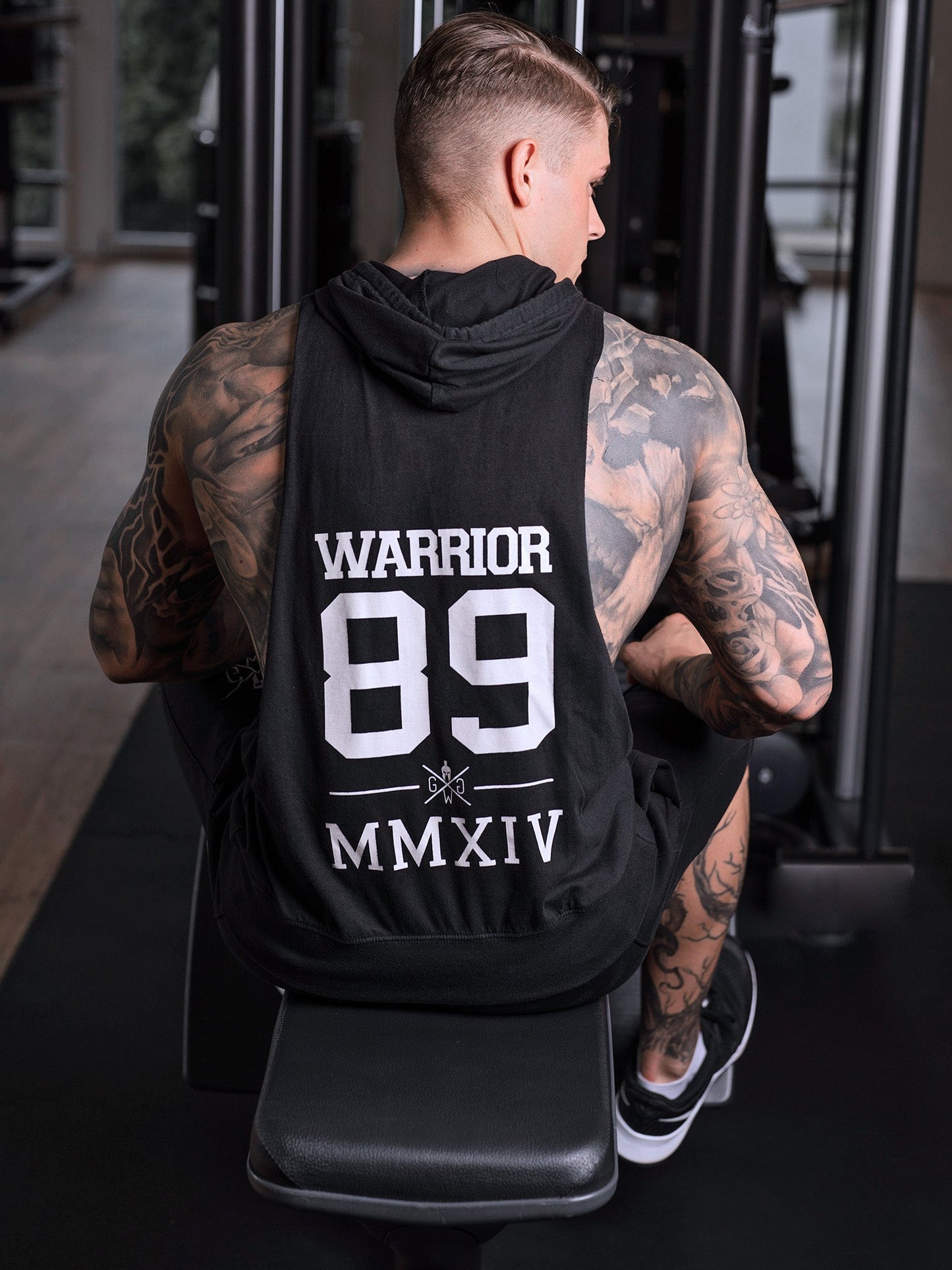 Back view of a muscular man wearing a black Gym Generation hoodie tank top with "Warrior 89 MMXIV" print, seated on a gym machine, ideal for gym, bodybuilding, and athletic training.