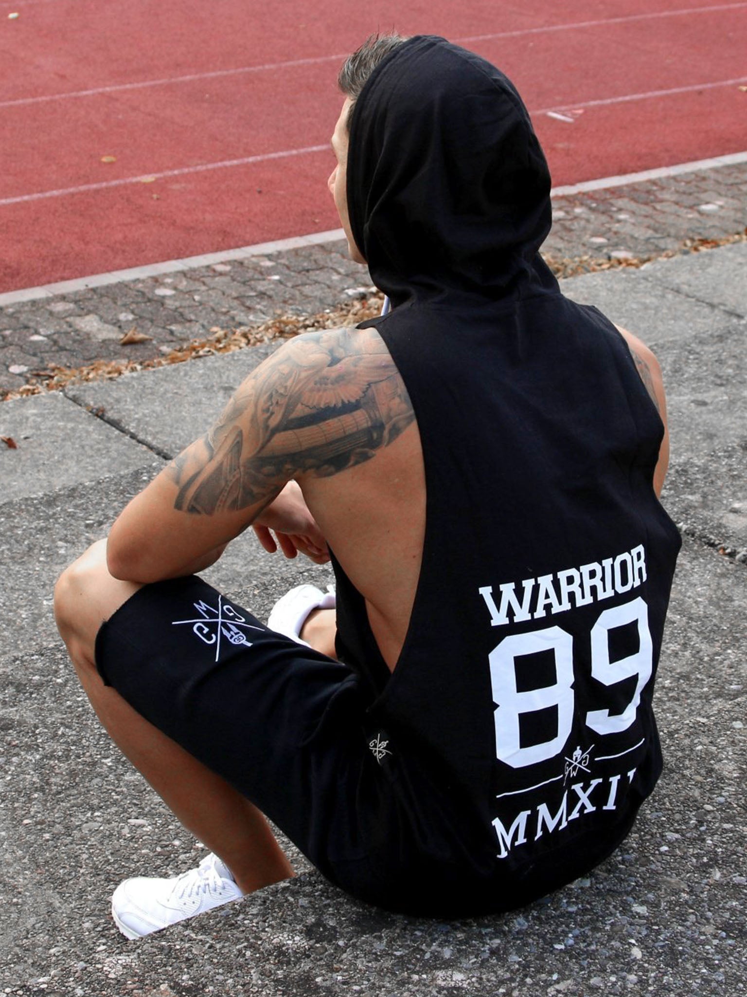 Man sitting on the ground wearing a black Gym Generation hoodie tank top with "Warrior 89 MMXIV" print and matching shorts, next to a running track, ideal for fitness, athletic training, and activewear style.